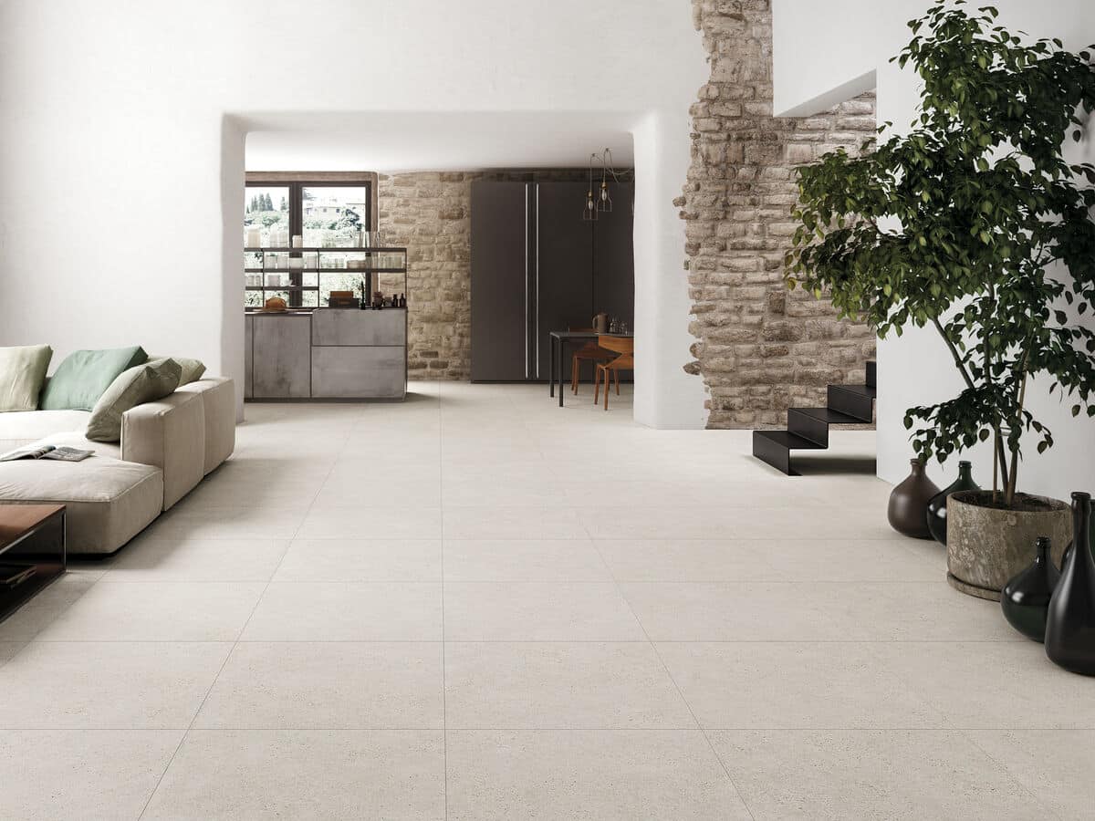 Ruime woonkamer met een landelijke vloer van lichte tegels, beige bank, groene planten en bakstenen muur gecombineerd met moderne inrichting.